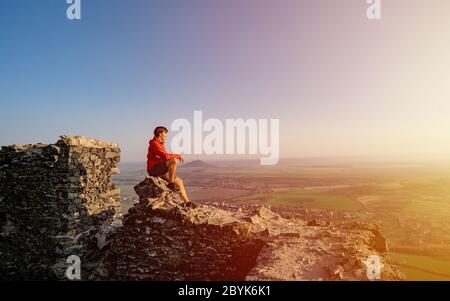 Homme professionnel assis sur les ruines du château vêtu d'une tenue active, reposant sur le sommet de la montagne et admirant les collines verdoyantes et les paysages à couper le souffle Banque D'Images