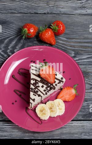 Cheesecake sur une assiette. Garni de fraises, bananes et chocolat. Sur les planches de pin brossé peintes en noir et blanc. Banque D'Images