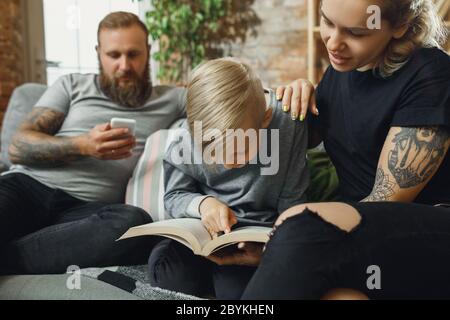 Famille heureuse à la maison passant du temps ensemble. S'amuser, avoir l'air gai et charmant. Mère, père et fils lisant le livre pendant les études à distance. Parents aidant. Enfance, concept de vie domestique. Banque D'Images