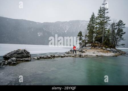 Homme en veste rouge regardant sur le lac gelé. Petite île avec arbres au lac Eibsee Bavaria Zugspitze. Belle journée d'hiver Banque D'Images