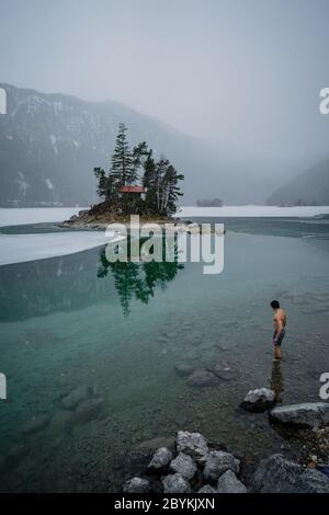 Homme en veste rouge regardant sur le lac gelé. Petite île avec arbres au lac Eibsee Bavaria Zugspitze. Belle journée d'hiver Banque D'Images