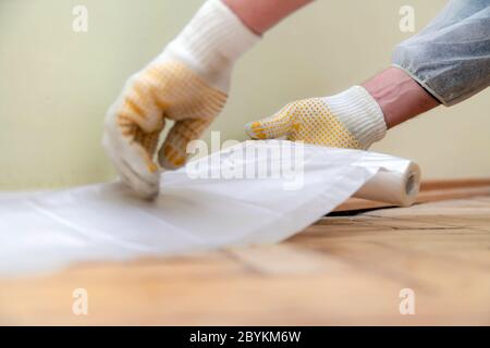 Travailleur avec des gants et des vêtements de travail de protection collant cellophane pour protéger les planchers en bois dur - préparant la salle pour la peinture Banque D'Images