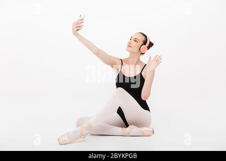 Image de la jeune femme caucasienne ballerine dans les écouteurs prenant photo selfie sur le téléphone portable isolé sur fond blanc de mur Banque D'Images