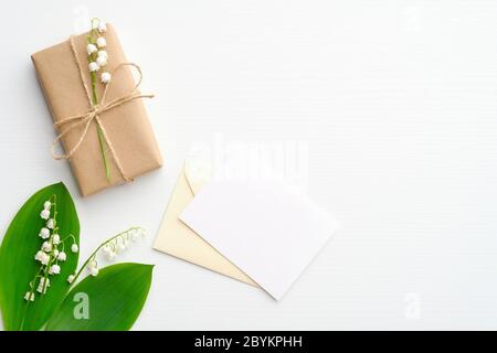 Boîte cadeau ou cadeau enveloppée de papier kraft, de fleurs de la vallée et de lettre romantique avec carte en papier vierge sur table blanche. Flat lay, vue de dessus. Banque D'Images