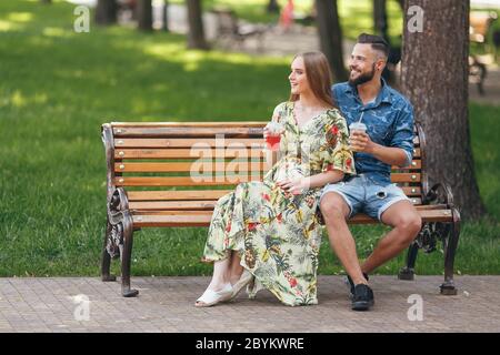 Jeune couple adolescent à la mode se reposant dans un parc de la ville avec des boissons assis sur un banc lors d'une journée ensoleillée d'été Banque D'Images