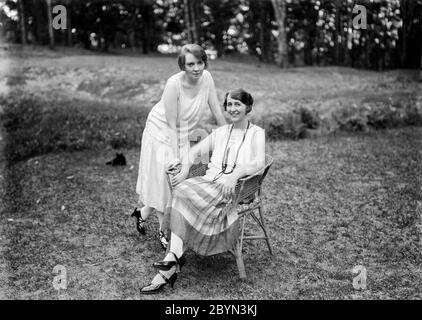 Photographie anglaise vintage des années 1920 en noir et blanc montrant une femme mûre, assise dans un jardin sur une chaise en osier, avec sa fille, une jeune femme, debout à côté d'elle, penchée sur la chaise. Les deux sont souriants et montrent la robe et la mode de l'époque. Banque D'Images