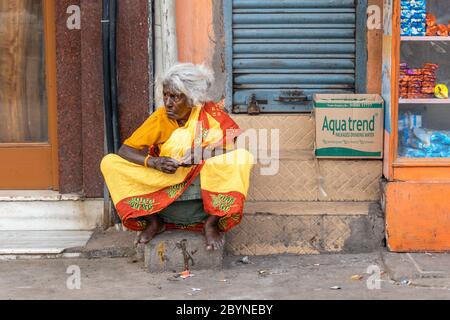 Chennai, Tamil Nadu, Inde - août 2018 : une femme indienne âgée portant un sari rouge et jaune coloré assis sur le trottoir. Banque D'Images