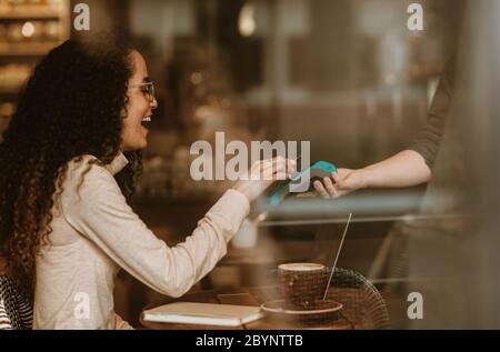 Femme souriante qui paie à l'aide de la technologie NFC dans un café pour payer sa facture. Une cliente qui paie sa facture à l'aide d'un smartphone et d'un lecteur électronique à c Banque D'Images