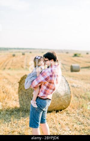 Joyeux jeune père joyeux, beau barbu homme en Jean et en chemise à carreaux, tenant les mains de sa jolie petite fille en robe, embrassant et Banque D'Images