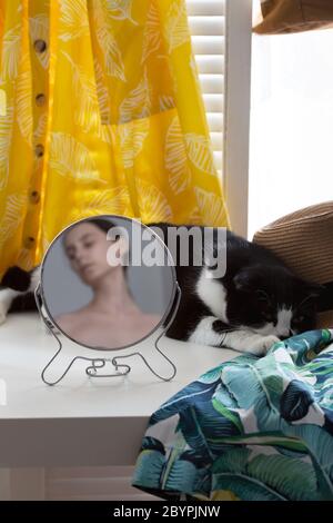 Sur la table près de la fenêtre dort chat autour des vêtements dans le miroir circulaire reflète la femme hors de focus Banque D'Images