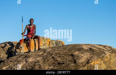 Le jeune guerrier Masai est assis sur une grande pierre en vêtements traditionnels avec une lance dans la savane contre un ciel bleu. Tanzanie, Afrique de l'est. Banque D'Images