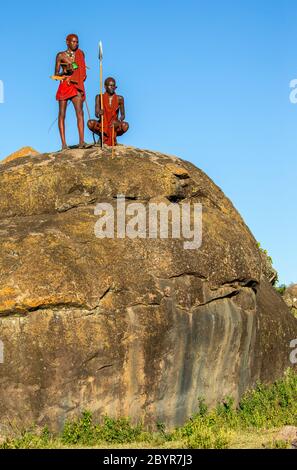 Deux jeunes guerriers Masai sur une grande pierre en vêtements traditionnels avec une lance dans la savane contre un ciel bleu. Tanzanie, Afrique de l'est, 12 août 2018 Banque D'Images