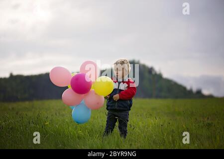 Petit enfant, mignon garçon sur un printemps froid venteux pluie jour, tenant des ballons colorés dans un champ, courir heureusement Banque D'Images