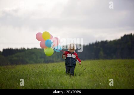 Petit enfant, mignon garçon sur un printemps froid venteux pluie jour, tenant des ballons colorés dans un champ, courir heureusement Banque D'Images