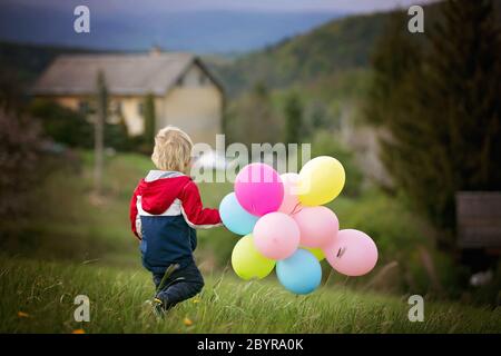 Petit enfant, mignon garçon sur un printemps froid venteux pluie jour, tenant des ballons colorés dans un champ, courir heureusement Banque D'Images