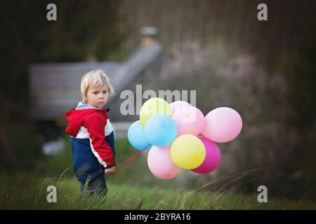 Petit enfant, mignon garçon sur un printemps froid venteux pluie jour, tenant des ballons colorés dans un champ, courir heureusement Banque D'Images