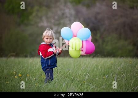 Petit enfant, mignon garçon sur un printemps froid venteux pluie jour, tenant des ballons colorés dans un champ, courir heureusement Banque D'Images