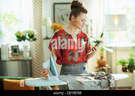 femme pensive élégante en blouse rouge et jupe crayon grise avec fer à repasser vérifier comment repasser correctement sur internet sur un smartphone dans le moderne ro de vie Banque D'Images