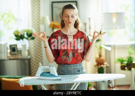 femme élégante et décontractée en blouse rouge et jupe crayon grise avec fer et planche à repasser faisant du yoga dans la maison moderne par temps ensoleillé. Banque D'Images