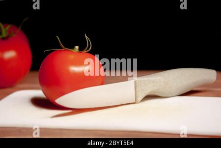Tomate rouge mûre avec couteau en céramique blanc et découpe de la table blanche sur fond noir Banque D'Images