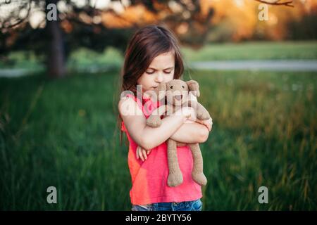 Triste contrarié caucasien fille embrassant jouet. L'enfant s'étreinte d'un ours en peluche dans le parc à l'extérieur. Enfant solitaire perdu dehors. Des problèmes d'enfance malheureux Banque D'Images