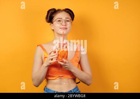 Une fille aux cheveux rouges en verres ronds et un haut orange est debout sur un fond jaune, souriant joli avec une limonade dans les mains Banque D'Images