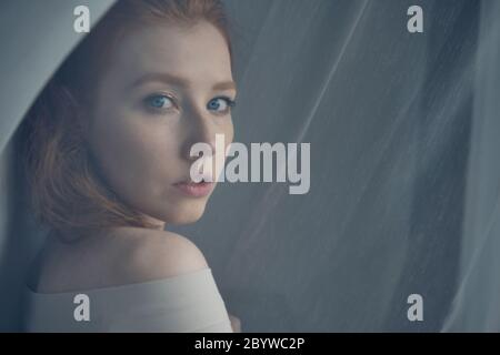 Une fille aux cheveux rouges avec des yeux bleus se tient au milieu d'un rideau translucide et regarde sur l'épaule, une vue à travers le rideau. Banque D'Images
