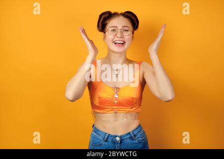 REDHEAD fille en verres ronds et un haut orange se tient sur un fond jaune, rires, élevant les paumes vers le haut Banque D'Images