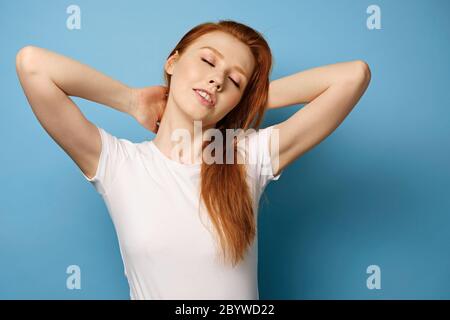 La jeune fille à cheveux rouges dans un T-shirt blanc se tient sur un fond bleu, avec ses mains derrière son cou et fermant ses yeux. Banque D'Images