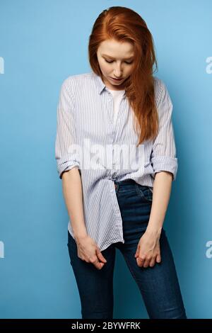 Une fille aux cheveux rouges dans une chemise rayée se tient sur un fond bleu et, avec sa tête courbée, tient le bord d'une chemise ample Banque D'Images