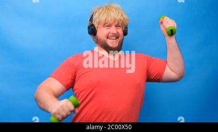 Drôle de gras homme freaky dans une perruque et un t-shirt rose avec des haltères faisant des exercices sur un fond bleu. Banque D'Images