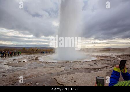 Personnes prenant des photos avec des téléphones cellulaires de la pittoresque Strokkur Geyser tout en éruptant, l'Islande automntime Banque D'Images