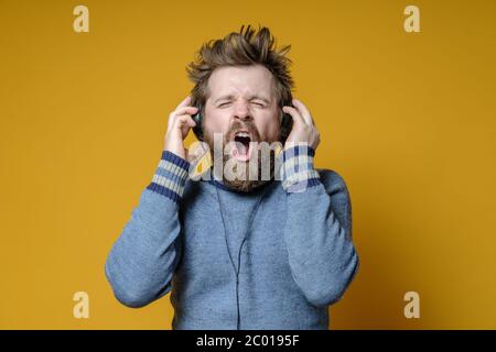 Un hipster satisfait dans un ancien pull écoute de la musique sur des écouteurs et chante fort, sur un fond jaune. Banque D'Images