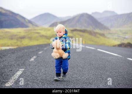 Un adorable petit garçon avec un ours en peluche à la main, qui court sur une route en Islande par temps de pluie Banque D'Images