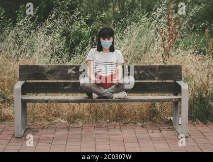 femme portant un masque médical lisant un livre assis sur un banc à l'extérieur Banque D'Images