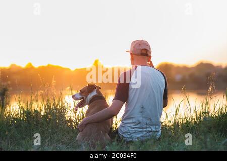 L'homme hopte son chien au coucher du soleil au bord du lac. Animaux de compagnie et amitié humaine, prendre soin, passer du temps ensemble. Banque D'Images