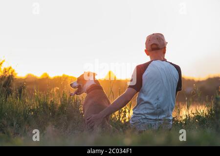 L'homme hopte son chien au coucher du soleil au bord du lac. Animaux de compagnie et amitié humaine, prendre soin, passer du temps ensemble. Banque D'Images