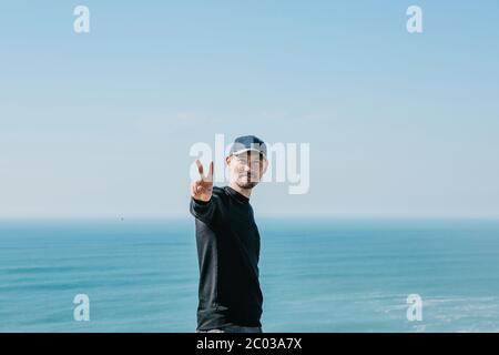 Un gars positif montre ses doigts un signe qui signifie la paix ou la victoire sur fond de mer ou d'océan. Banque D'Images