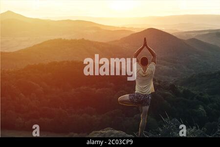 Jeune homme faisant du yoga sur le pic de montagne. Pratique le yoga et médite sur la montagne. Paysage vallonné loin des gens au coucher ou au lever du soleil Banque D'Images