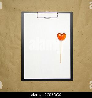 La tablette avec un clip pour le clip de papier avec une feuille blanche a4 repose sur l'arrière-plan du papier brun froissé. Sur le dessus se trouve un lollipop de caramel de rouge orange en forme de coeur. Place pour le texte et la mise en page pour la conception. Banque D'Images