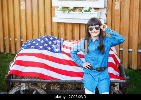 La fille se tient dans la cour, vêtue d'une robe en denim et tenant le drapeau des Etats-Unis Banque D'Images