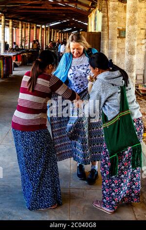 Un touriste essaie un long birman traditionnel (Sarong) aidé par deux femmes locales, le marché de Thaung Thut, le lac Inle, l'État Shan, Myanmar. Banque D'Images