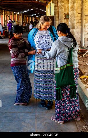 Un touriste essaie un long birman traditionnel (Sarong) aidé par deux femmes locales, le marché de Thaung Thut, le lac Inle, l'État Shan, Myanmar. Banque D'Images