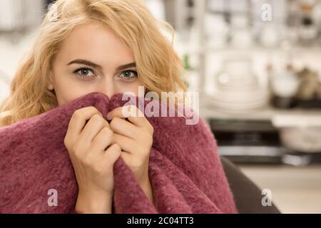 Gros plan d'une femme blonde heureuse qui se retrouve devant l'appareil photo, enveloppée d'une couverture en laine chaude dissimulant son visage, se détendant à la maison dans un espace d'imitation Banque D'Images