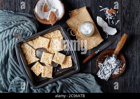 barres de crack de noix de coco sans cuisson dans un plat de cuisson sur une table rustique en bois avec des ingrédients et de l'huile de coco biologique dans un pot, vue horizontale d'en haut, fl Banque D'Images