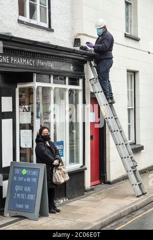 Un ingénieur de téléphone portant un casque, un masque et des gants alors qu'il travaille à l'extérieur d'une pharmacie, à côté d'un client masqué de même type, Presteigne, au pays de Galles, au Royaume-Uni Banque D'Images