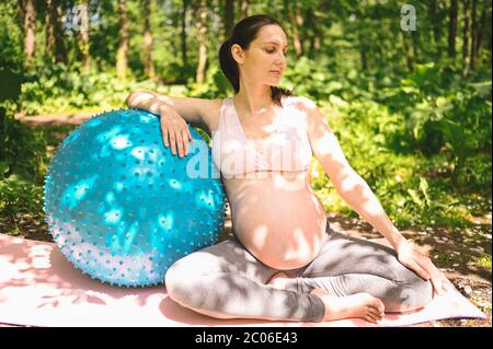 Belle jeune femme enceinte faisant de l'exercice avec la forme physique pilates Blue ball dans le parc extérieur. Asseyez-vous et détendez-vous sur un tapis de yoga rose. Un avenir actif Banque D'Images