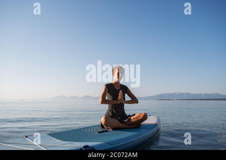 Jeune femme bronzée assise en position lotus avec ses mains jointes devant sa poitrine, méditant sur le plateau flottant sur l'eau de mer calme matin. Banque D'Images