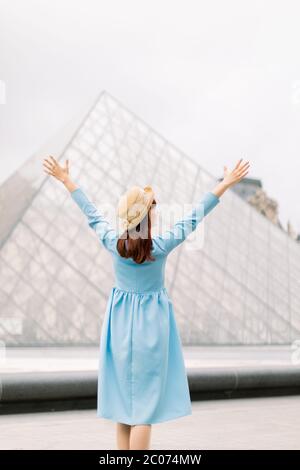 PARIS, FRANCE - 17 septembre 2019 : bonne et heureuse jeune fille en chapeau et robe bleue debout près de la pyramide de verre dans le musée du Louvre avec les bras vers le haut et Banque D'Images