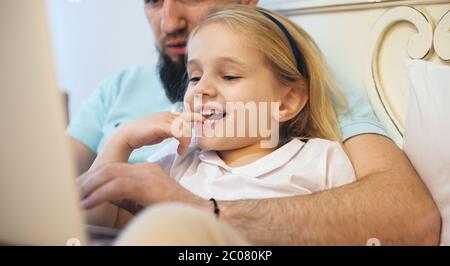 Charmante blonde fille passant du temps avec son père barbu allongé sur le canapé et lisant quelque chose Banque D'Images